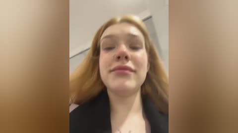 Media: Video of a fair-skinned woman with long, auburn hair, wearing a black top, looking down with a neutral expression, blurred background.