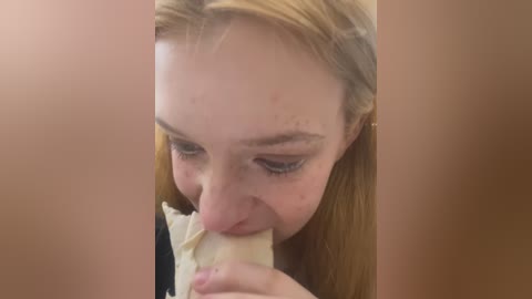 Media: Video of a young Caucasian woman with light skin, straight blonde hair, and freckles, licking a piece of cream cheese off a sandwich, set against a plain brown background.