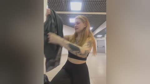 Media: Video of a blonde woman in a gym, wearing a yellow crop top and black leggings, holding a black jacket, with a tattooed arm, in a modern, bright gym with grid ceiling and white walls.