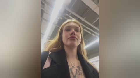 Media: Video of a young woman with long blonde hair, wearing a black coat, standing indoors with fluorescent lighting. She has a tattoo on her chest and looks confident.