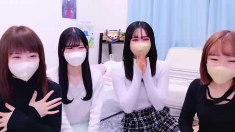 Media: Video of four East Asian women in masks, wearing white tops and plaid skirts, sitting in a brightly lit room with blue curtains and a white couch.