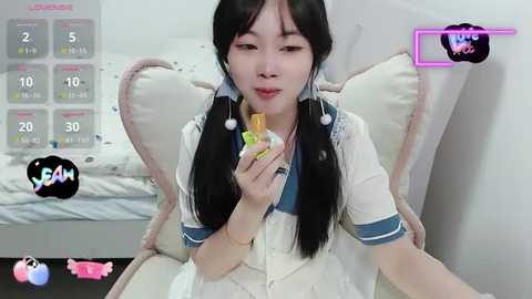 Media: Video of an Asian woman with long black hair in pigtails, wearing a white sailor outfit, holding a colorful sex toy, against a white background.