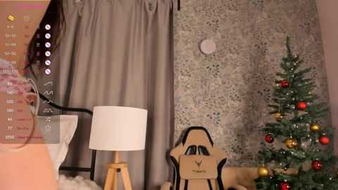 Video of a cozy room with a grey curtain, a white lamp, a wooden chair, and a small, decorated Christmas tree with red and gold ornaments.