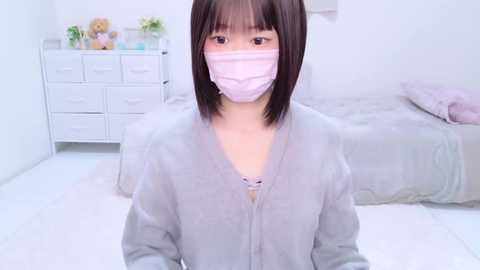 Media: A video of a young Asian woman with straight black hair, wearing a light pink face mask and gray cardigan, standing in a bright, minimalist bedroom with white walls, a bed, and a dresser.