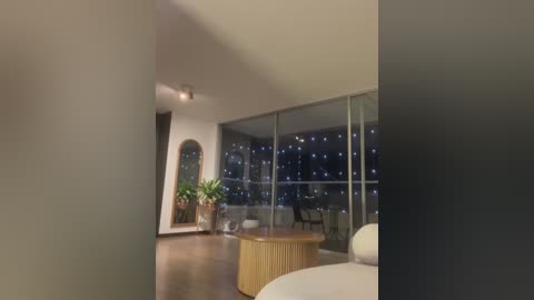 Video of a modern, dimly lit living room with a large glass window revealing a nighttime cityscape. A round, wooden coffee table with potted plants sits on a polished hardwood floor.