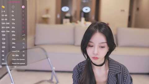 Media: Video of a young Asian woman with long black hair, wearing a black and white striped blazer, sitting on a light-colored couch in a modern living room.