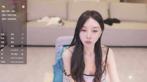 Media: Video of an East Asian woman with long black hair, wearing a white camisole, sitting on a chair in a modern, beige sofa-laden living room.