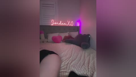 A dimly lit bedroom video features a person in black underwear lying on a bed with beige bedding and plush toys, illuminated by pink and purple neon lights.