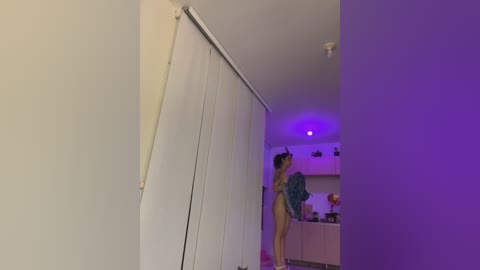 Media: A video of a slender, nude woman with long brown hair, wearing a blue shirt, standing in a dimly lit, modern kitchen with white cabinets and purple lighting.