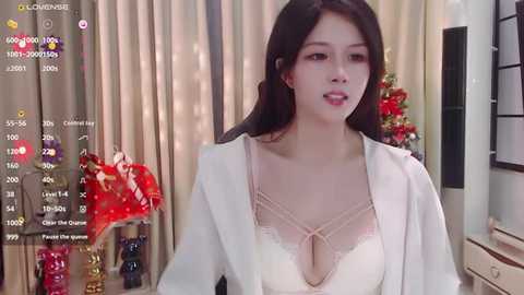 Media: Video of an East Asian woman with long black hair, wearing a white blazer over a revealing lace bra, in a festive room with Christmas decorations, a camera overlay on the image.