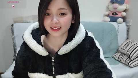 Media: Video of an Asian woman with straight black hair, wearing a black and white fuzzy hoodie, sitting on a striped couch in a cozy room with plush toys.