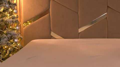 Media: Video of a luxurious, modern bedroom with a beige upholstered headboard, warm ambient lighting, and a festive, decorated Christmas tree on the left, adorned with golden baubles and fairy lights.