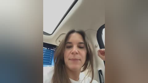 Media: Video of a young woman with long brown hair, wearing a white top, looking relaxed with eyes closed in a car's backseat, with a window and ceiling light visible.