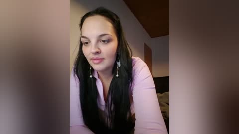 Media: Video of a young Asian woman with long black hair, wearing a light pink blouse, leaning forward, with a neutral expression, in a dimly lit, modern bedroom with a wooden headboard.