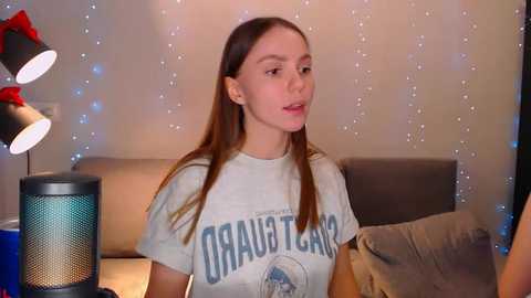 Media: Video of a young woman with long brown hair, wearing a gray \"Orange County\" t-shirt, sitting indoors with a modern, cozy atmosphere featuring a gray sofa, blue lamp, and string lights.