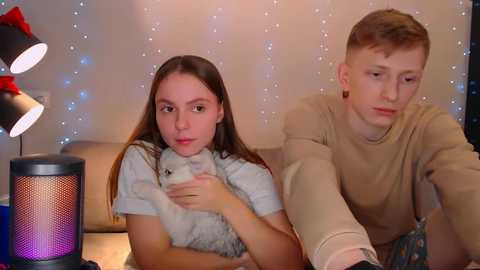 Media: Video of a young Caucasian couple, a girl with long brown hair and a boy with short blonde hair, cuddling a small white cat in a cozy living room with twinkling fairy lights and modern decor.