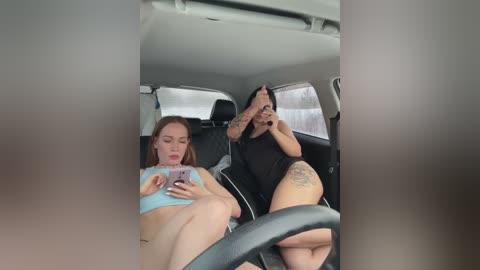 Media: Video of two women in a car, one wearing a light blue crop top and the other a black shirt, both with tattoos, taking selfies.