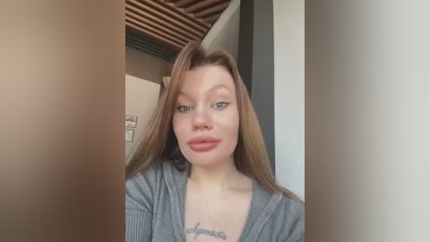 Media: Video of a young, light-skinned woman with straight, shoulder-length auburn hair, wearing a grey ribbed cardigan, standing indoors near a window with wooden blinds. She has a tattoo on her chest and is smiling slightly.