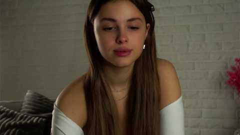 Media: Video of a young woman with long brown hair, fair skin, wearing a white off-shoulder top, and a gold necklace, standing against a white brick wall with pink flowers in the background.