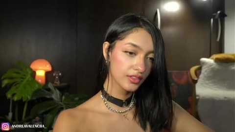 Media: Video of a young Asian woman with long black hair and light skin, wearing a black choker, seated in a dimly lit room with a glowing mushroom lamp and potted plants.