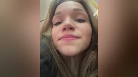 A close-up video of a smiling young woman with light skin and long blonde hair, wearing a dark jacket, taken from an upward angle.