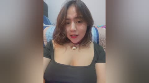 Media: A video of an Asian woman with shoulder-length brown hair, wearing a black top with a bell choker, sitting in a bedroom with a bed and colorful blankets in the background.