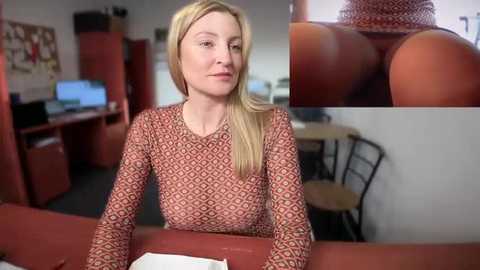 Media: Video of a blonde woman in a patterned brown top, sitting at a red desk in an office. Close-up inset of a hand holding a hotdog bun.