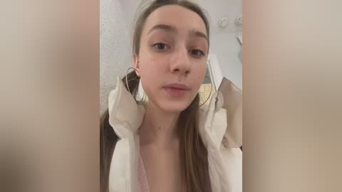 Media: Video of a young woman with fair skin and straight brown hair, wearing a white blouse, standing in a room with a white ceiling and a white door.