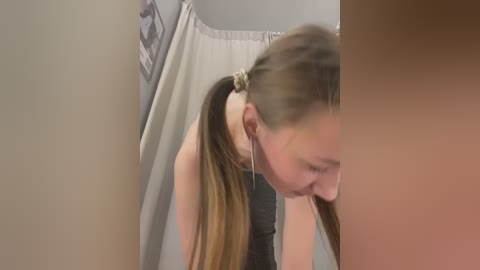 Media: Video of a young woman with long, light brown hair in a ponytail, wearing a grey tank top, leaning forward in a public restroom stall, earbuds in, with a beige curtain in the background.