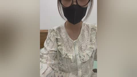 Media: Video of an Asian woman with short, dark hair, wearing a black face mask, black-rimmed glasses, and a white lace blouse with floral patterns. She is indoors, with a wooden wall and a blurred background.