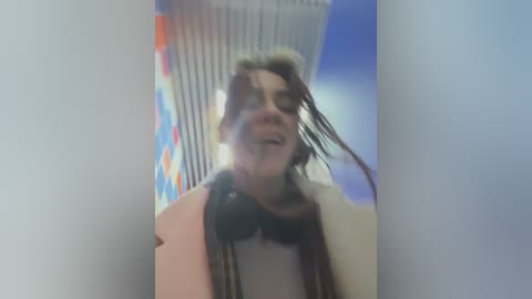 Media: A blurry, out-of-focus video of a person with short, curly hair, wearing a beige coat and black headphones, standing in front of a vending machine with colorful, vertical stripes.