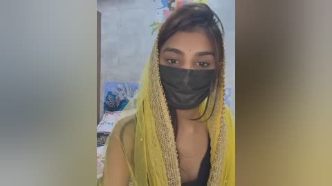 Media: Video of a young South Asian woman wearing a yellow dupatta, black face mask, and brown hair, indoors with a blurred background of a cluttered room.