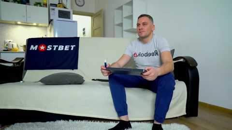 Media: Video of a man in a gray T-shirt and blue pants, sitting on a white couch in a modern kitchen, holding a tablet.