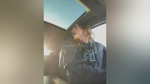 Media: Video of a young woman with long, wavy blonde hair, wearing a dark hoodie with white lettering, gazing out the car window at a sunny day.