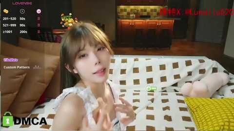 Media: Video of an Asian woman with straight brown hair, wearing a white lace dress, lying on a bed with a brown and white patterned duvet. A toy bear and a green remote control are on the bed. Background shows a modern kitchen with wooden cabinets.