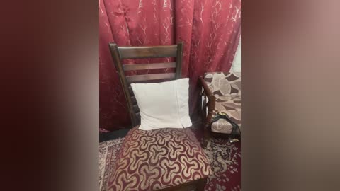 Media: Video of a wooden chair with a white pillow and a patterned red and brown throw, set against a maroon, floral-patterned curtain backdrop in a cozy, vintage room.