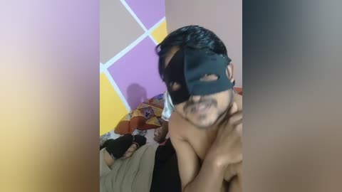 Media: Video of a young, shirtless Indian man with a black mask, standing in a room with a bed and colorful, geometric-patterned wall.