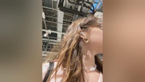 Media: Video of a young woman with long, wavy brown hair, wearing a black strapless top and gold hoop earrings, against an industrial background with metal beams and pipes.