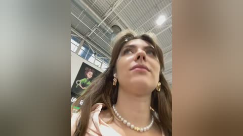 Media: Video of a young woman with long brown hair, wearing a white top and pearl necklace, gazing upwards in a high-ceilinged room with industrial lighting and blurred background.