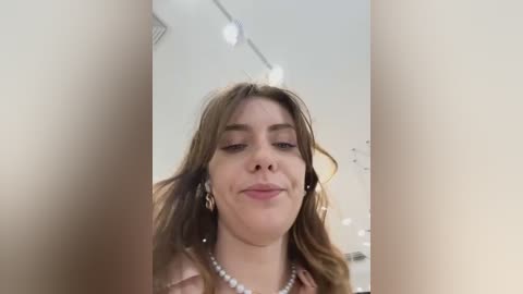 Media: A video of a smiling, light-skinned woman with wavy brown hair, wearing a pearl necklace, standing in a blurred, indoor setting with a white ceiling and a light-colored wall.