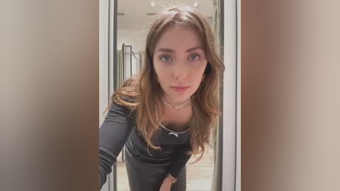 Media: Video of a young woman with light skin, wavy brown hair, and blue eyes, wearing a black long-sleeve top, standing in a modern hallway with beige walls and a glass door.