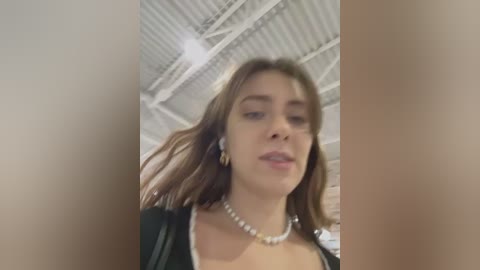 Media: Video of a young Caucasian woman with light skin, brown hair, and a slim physique, wearing a black jacket and pearl necklace, standing in an industrial, white-beamed room.