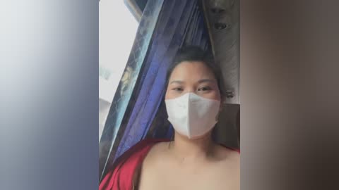 Video of a woman with light skin, wearing a white surgical mask, and a red top, sitting in a dimly lit room with blue curtains and a wooden door.