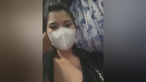 Video of a woman with medium brown skin, wearing a white face mask, black top, and a patterned curtain in the background.