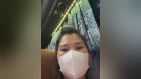 Media: Video of a woman wearing a white face mask, sitting in a bus with blue and green patterned curtains, yellow string lights, and a brown seat visible in the background.