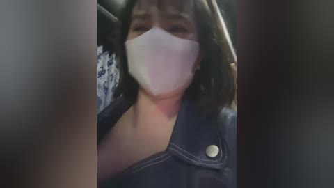 Media: A video shows a woman with light skin, shoulder-length dark hair, and a white mask, wearing a dark leather jacket. She is seated in a dimly lit interior, possibly a car.