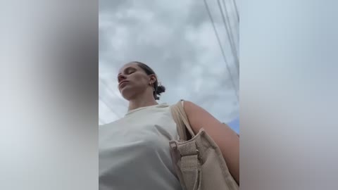 Media: A video of a tall woman with a fair complexion, wearing a beige sleeveless top, standing against a cloudy sky. She carries a beige handbag. The image is slightly blurry, giving a dreamy, ethereal feel.