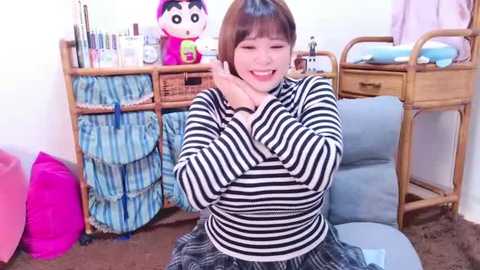 Media: Video of a smiling Japanese woman with straight brown hair, wearing a black-and-white striped top and a plaid skirt, sitting on a blue cushion in a cluttered room with wicker furniture, toys, and pink bags.