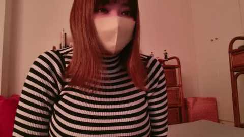 Media: Video of a young woman with long, straight brown hair, wearing a black and white striped sweater, a face mask, and standing in a dimly lit, cluttered room with red furniture and a wicker chair.