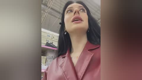 Media: Video of a woman with long black hair, fair skin, and wearing a red leather jacket, standing in a warehouse with shelving units and a metallic ceiling.